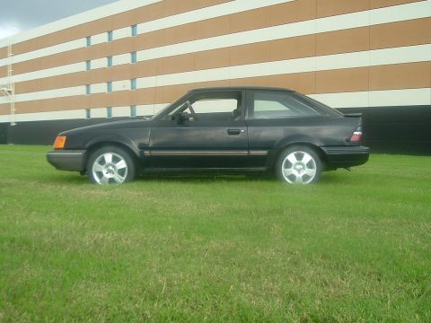 1989 Ford Escort - This dark-colored 1989 Ford Escort coupe is seen from the side, parked on a grassy area in front of a building.
