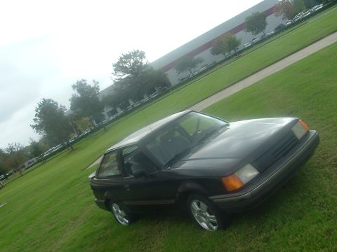 1989 Ford Escort - A dark hatchback is parked on a grassy area with a large industrial building in the background.