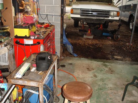 1989 Ford Bronco - A white Ford F-Series truck is undergoing repairs in a cluttered garage, with tools and equipment scattered around.