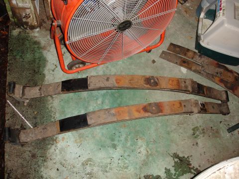 1989 Ford Bronco - Leaf springs, likely for a vehicle suspension, are shown on a workshop floor with a large industrial fan in the background.