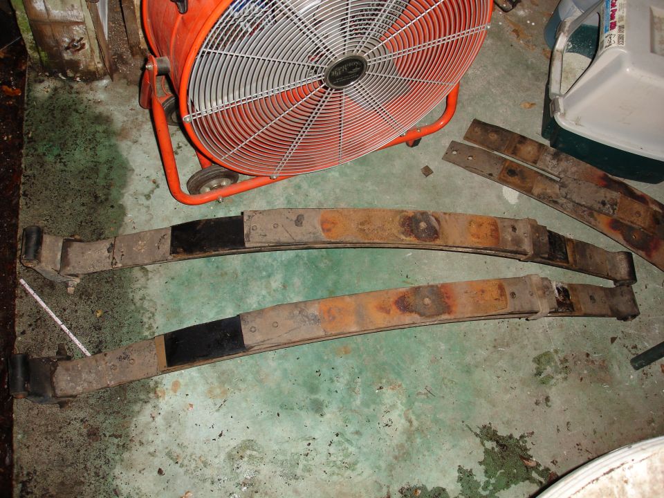 Two rusty leaf springs with black-painted ends sit on a concrete floor next to a large orange fan