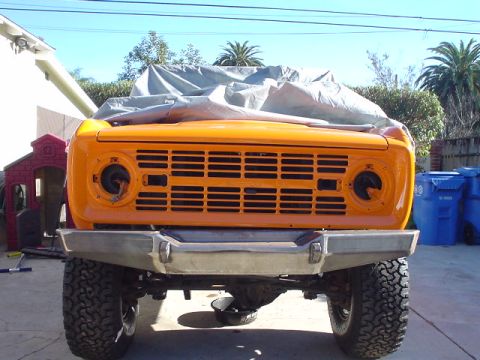 1977 Ford Bronco - The front grille and bumper of an orange 1977 Ford Bronco are visible, with a tarp covering the upper portion of the vehicle.