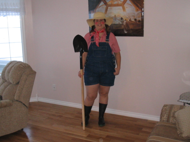 Person in denim overalls, pink shirt, red bandana, and straw hat holding a shovel