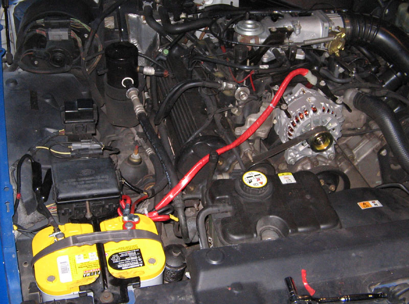 Engine bay with yellow Optima battery, red battery cable, alternator, and coolant reservoir