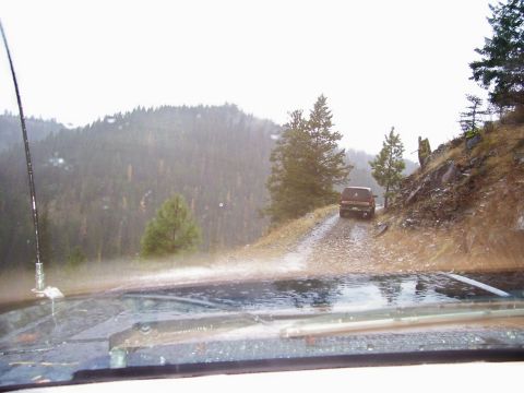1991 Ford Bronco - Two SUVs navigate a wet, mountainous dirt road in the rain.