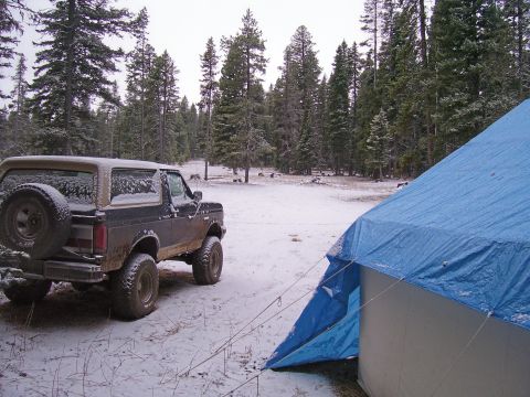 1991 Ford Bronco - A Ford Bronco sits in a snowy clearing surrounded by pine trees, with a blue tent set up nearby.