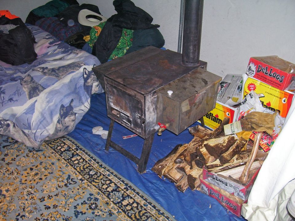 Wood stove with attached water tank and chimney pipe, next to firewood and boxes of "Del Logs"
