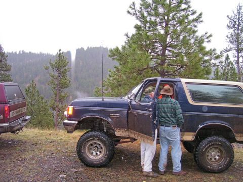 1991 Ford Bronco - A dark blue 1991 Ford Bronco with a tan top is parked on a dirt road with another vehicle visible to the left.