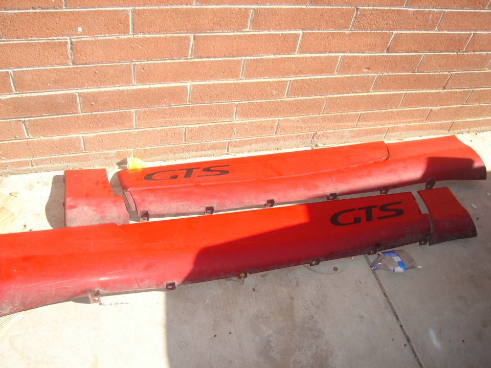 Two red car side skirts with black GTS lettering are laid out on concrete in front of a brick wall