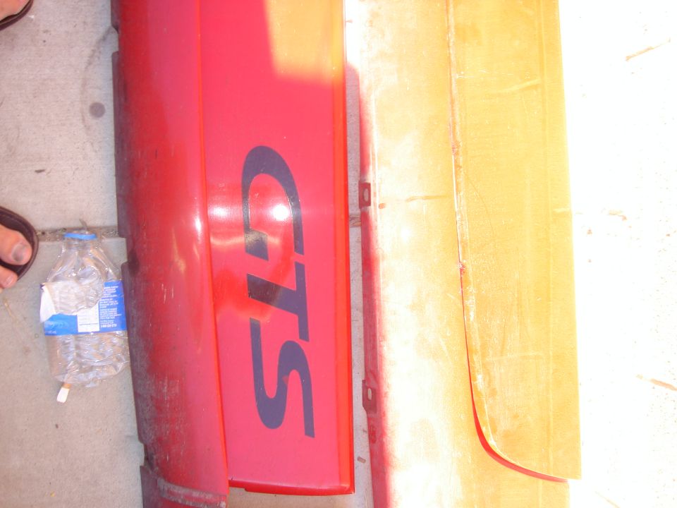 Red car bumper with dark blue GTS lettering, next to a yellowed panel. Water bottle on ground