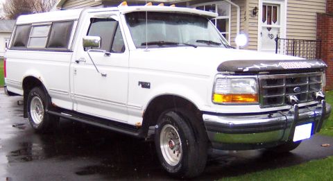 1978 Ford Bronco - A white pickup truck with a camper shell is parked on a wet driveway in front of a house.