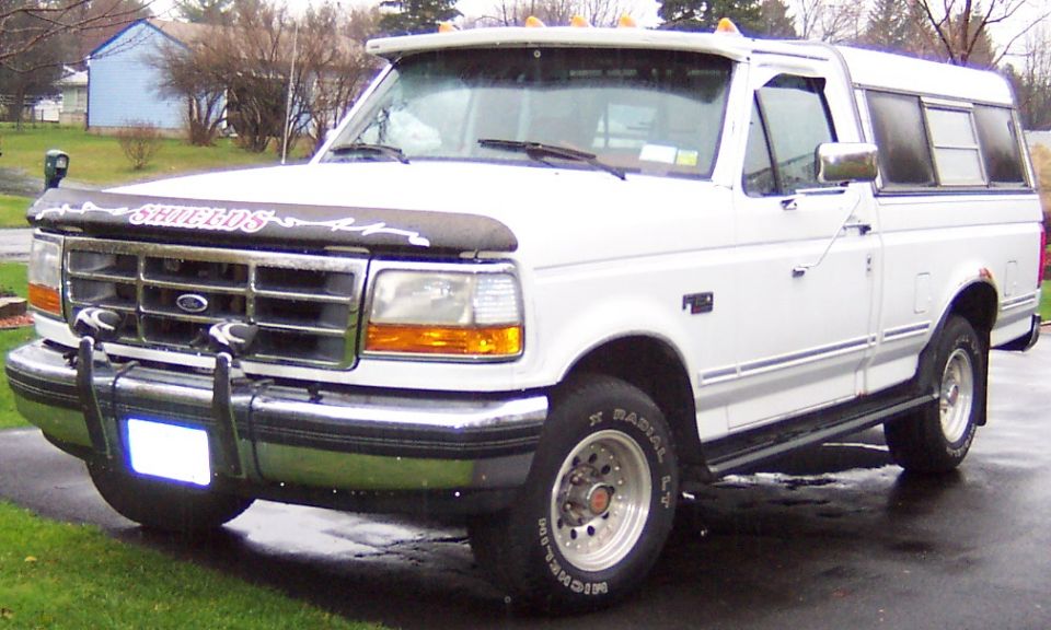 White Ford pickup truck with a camper shell, front-mounted bug deflector, and aftermarket lights