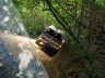 1989 GMC Jimmy - An off-road vehicle tackles a challenging muddy incline surrounded by dense forest foliage.