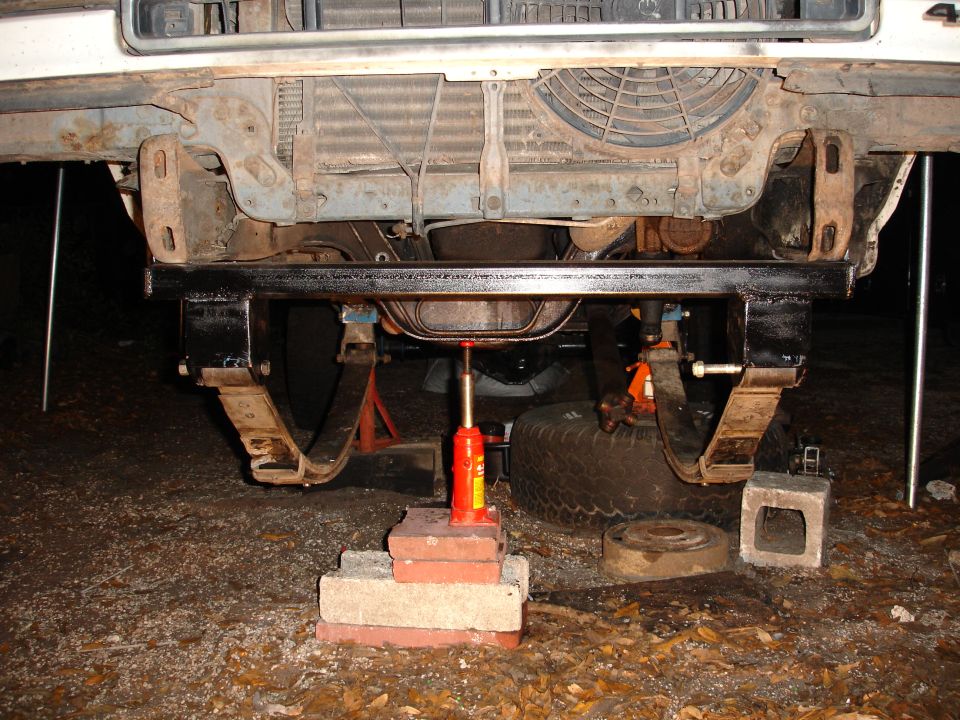 Front underside of a white vehicle being lifted by a red hydraulic jack on bricks, with leaf springs and a fan visible
