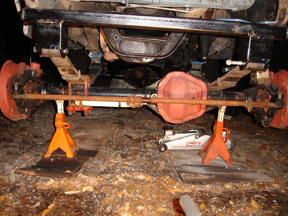 Vehicle rear axle assembly with leaf springs and brake drums, supported by jack stands. A Pro-Lift jack is visible