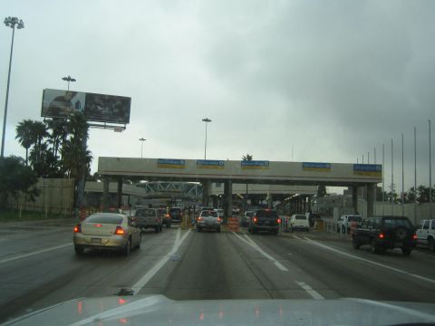 1990 Ford Bronco - Dashboard view of cars queued in lanes approaching a multi-booth border or toll crossing under a gray sky.