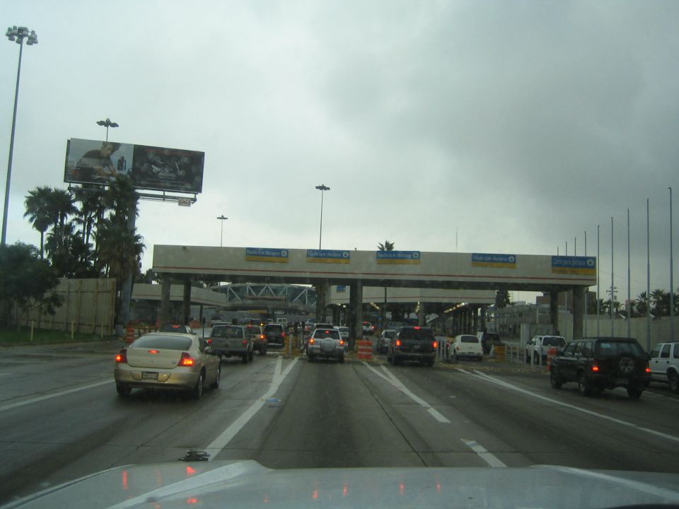 Cars queued at a multi-lane border or toll crossing under an overcast sky, dash view