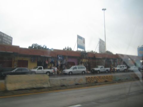 1990 Ford Bronco - row of open-air roadside market stalls under a red awning, with pickup trucks and minivans parked in front, shot through a car window.