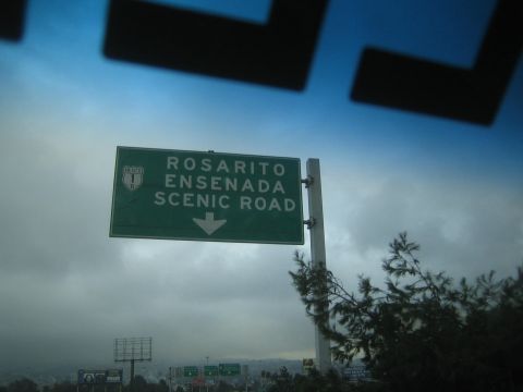 1990 Ford Bronco - green overhead highway sign reading 'Rosarito Ensenada Scenic Road' with a downward arrow against a cloudy sky.