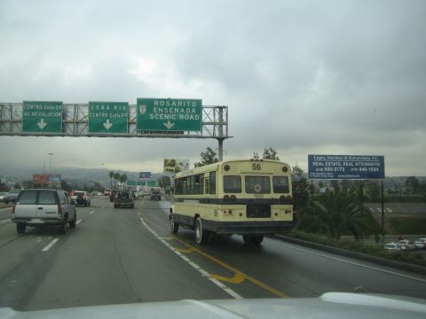 1990 Ford Bronco - yellow short bus numbered 56 travels a wet multilane highway beneath green Spanish-language overhead exit signs on an overcast day.