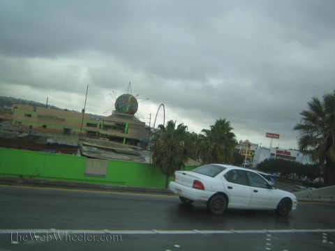 1990 Ford Bronco - white four-door sedan drives a wet road in front of a green-walled building topped with a large decorative globe sculpture.
