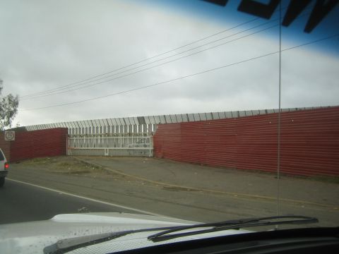 1990 Ford Bronco - long red corrugated-metal fence with a white gate runs beside a road, seen over a vehicle hood and windshield wiper.