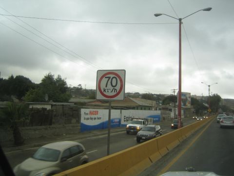 1990 Ford Bronco - round 70 km/h speed limit sign stands beside a busy multilane highway with passing cars and SUVs under an overcast sky.