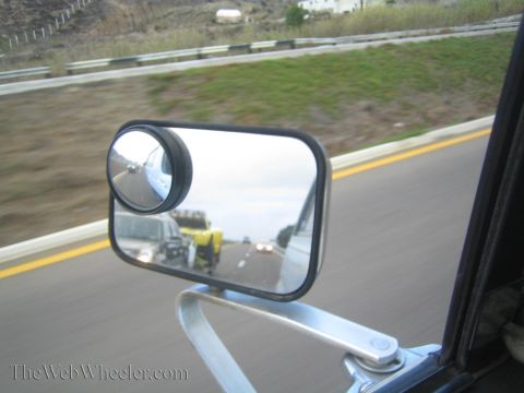 1990 Ford Bronco - round convex blind-spot mirror mounted on a chrome side mirror reflects traffic and a flatbed trailer on a coastal highway.