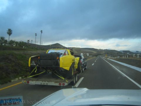 1990 Ford Bronco - yellow tube-frame off-road race buggy sits on a flatbed trailer towed by a dark truck along a coastal highway under stormy clouds.