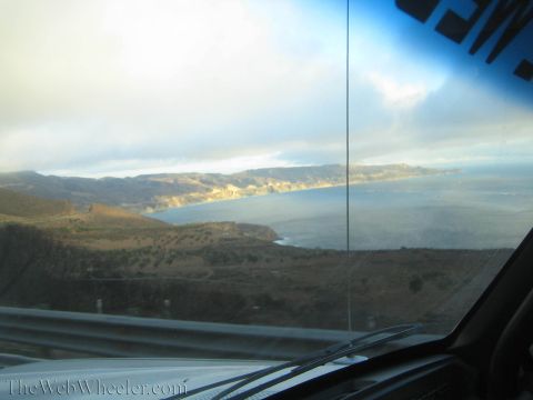 1990 Ford Bronco - wide coastal bay backed by hills, viewed through a car windshield with sun glare and dashboard reflections.