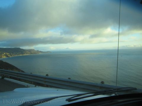 1990 Ford Bronco - calm ocean and rugged coastline stretch toward the horizon under broken clouds, seen over a car hood and windshield wiper.