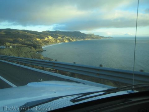 1990 Ford Bronco - Sunlit coastal cliffs curve along a bay beside a highway guardrail, viewed over a car hood under a partly cloudy sky.