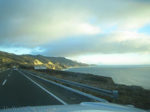 1990 Ford Bronco - two-lane coastal highway curves along sunlit cliffs above the ocean, with a small roadside sign and a distant car ahead.