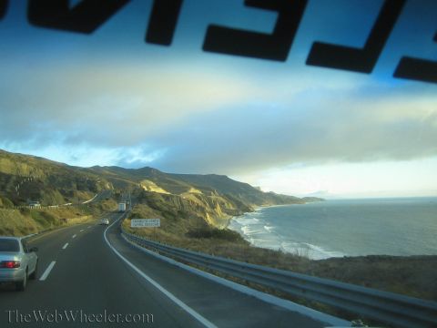 1990 Ford Bronco - coastal highway curves past sunlit hills and surf along the ocean, with a silver car alongside, seen through a windshield.