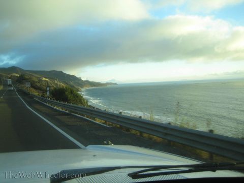 1990 Ford Bronco - sweeping ocean bay and distant headland line a guardrailed highway under heavy clouds, viewed over a car hood and wiper.