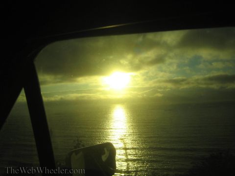 1990 Ford Bronco - golden sun sets over the ocean, its light reflecting on the water, framed by the dark interior pillar of a car window.