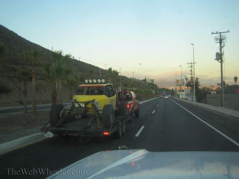 1990 Ford Bronco - yellow off-road race car on a flatbed trailer is towed by a red truck along a dusk highway lined with palms and hills.