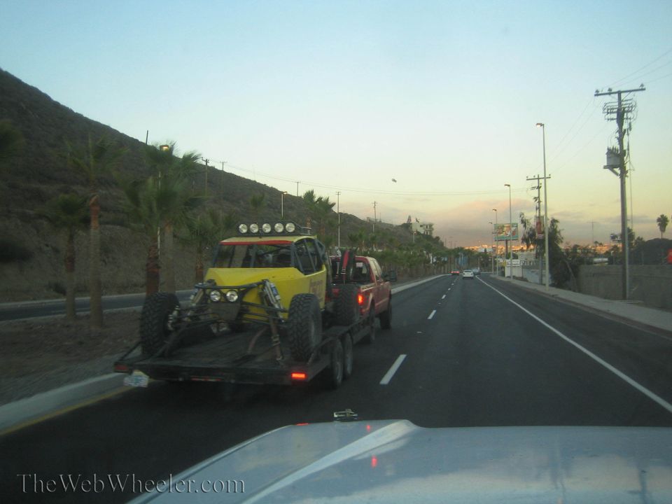 Yellow off-road race car on a flatbed trailer towed at dusk near palms