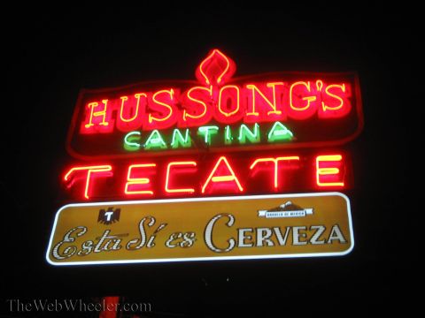 1990 Ford Bronco - glowing red and green neon sign reads 'Hussong's Cantina' and 'Tecate - Esta Si es Cerveza' against a black night sky.