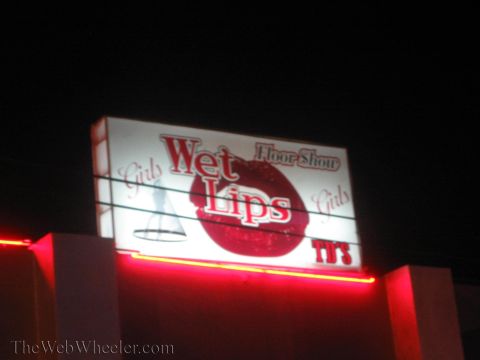 1990 Ford Bronco - backlit sign reading 'Wet Lips' with 'Floor Show' and 'Girls' text glows red and white against a dark night sky.