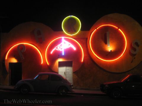 1990 Ford Bronco - building facade with large neon letters spelling 'PARIS' glows orange and pink at night, with an old VW Beetle parked at the curb.