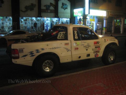1990 Ford Bronco - white Ford Ranger off-road race truck marked '226' with sponsor decals and a rear spare sits on a street at night.