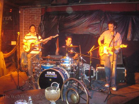1990 Ford Bronco - three-piece band plays on a small stage, with two guitarists and a drummer, a cocktail glass in the foreground.