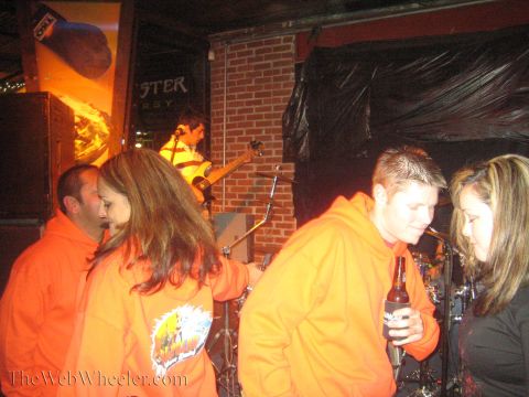 1990 Ford Bronco - crowd in matching orange hoodies stands near a brick-walled stage where a guitarist performs at a bar.
