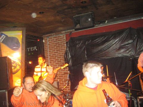 1990 Ford Bronco - People in orange hoodies fill a dim bar in front of a brick stage where a man plays guitar under a speaker.