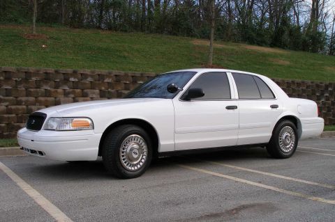 2001 Ford Crown Victoria - A white 2001 Ford Crown Victoria sedan is parked in a lot with a brick wall and trees in the background.