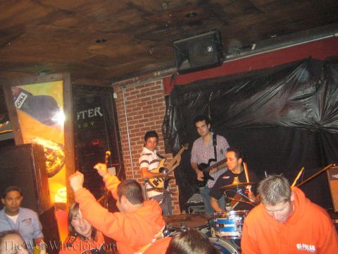 1990 Ford Bronco - cheering crowd in orange hoodies raises fists in front of a live band performing on a small brick-backed stage.