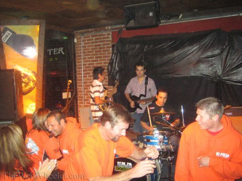 1990 Ford Bronco - group in orange shirts gathers around a bar table while a guitarist and drummer perform on the brick stage behind.