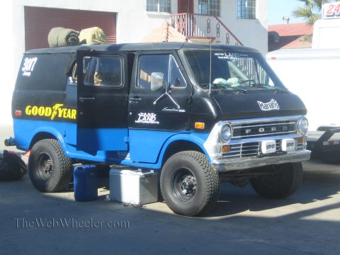 1990 Ford Bronco - lifted blue and black Ford 4x4 van with Goodyear and sponsor decals sits parked on big off-road tires outside a building.