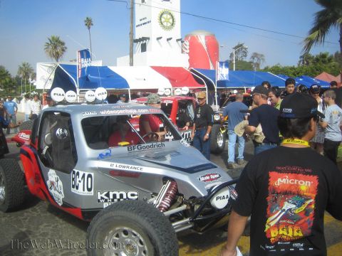 1990 Ford Bronco - silver off-road race buggy numbered 'BC-10' with BFGoodrich and KMC decals sits amid crowds and blue-striped tents at an event.